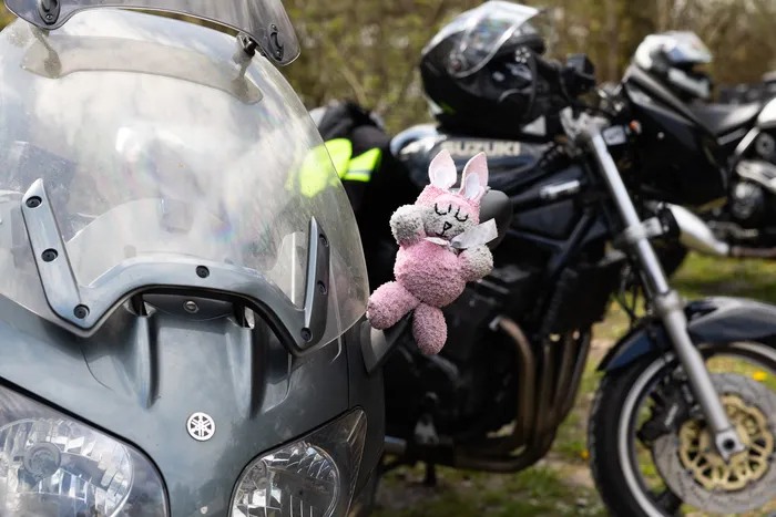 Ein rosa Stofftier-Hase mit Schleife ist an der Windschutzscheibe eines Motorrads befestigt, im Hintergrund weitere Motorräder und Motorradfahrer.