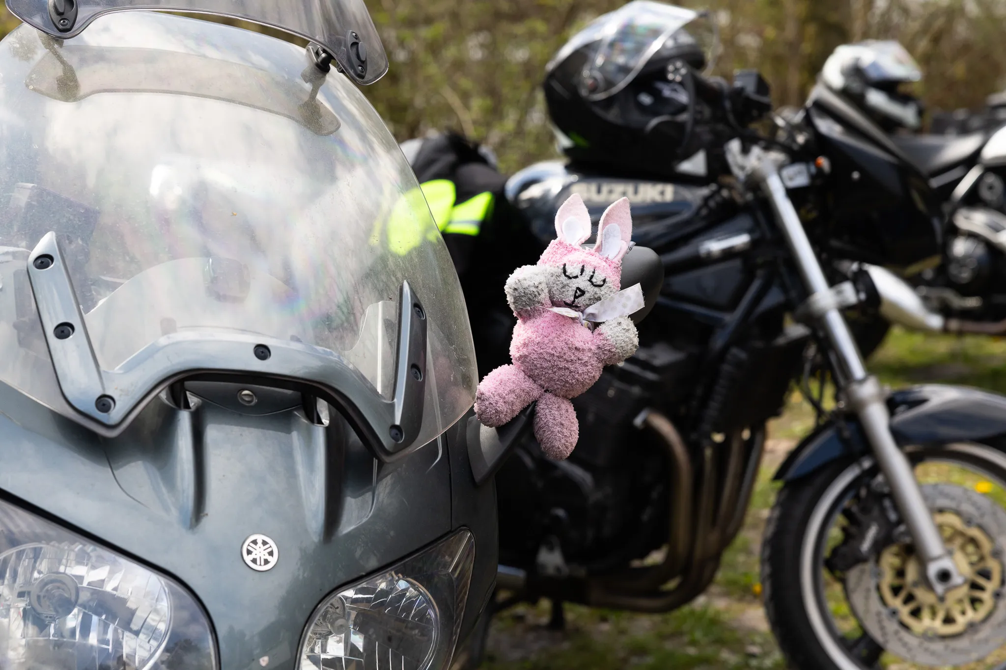 Ein rosa Stofftier-Hase mit Schleife ist an der Windschutzscheibe eines Motorrads befestigt, im Hintergrund weitere Motorräder und Motorradfahrer.