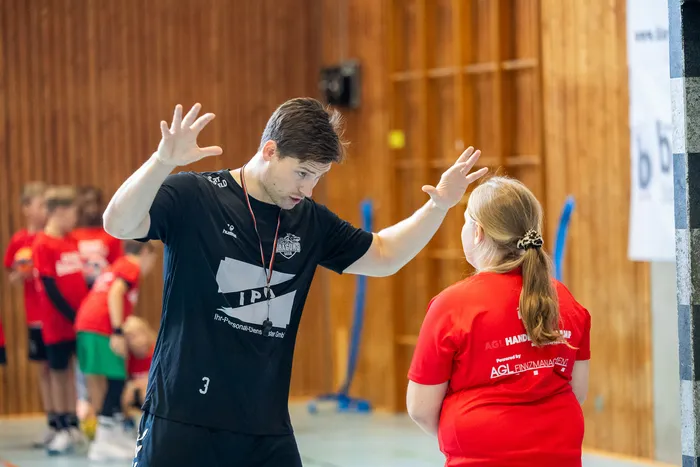 Ein Mann in einem schwarzen T-Shirt gestikuliert mit erhobenen Händen gegenüber einer Person in einem roten T-Shirt in einer Sporthalle.