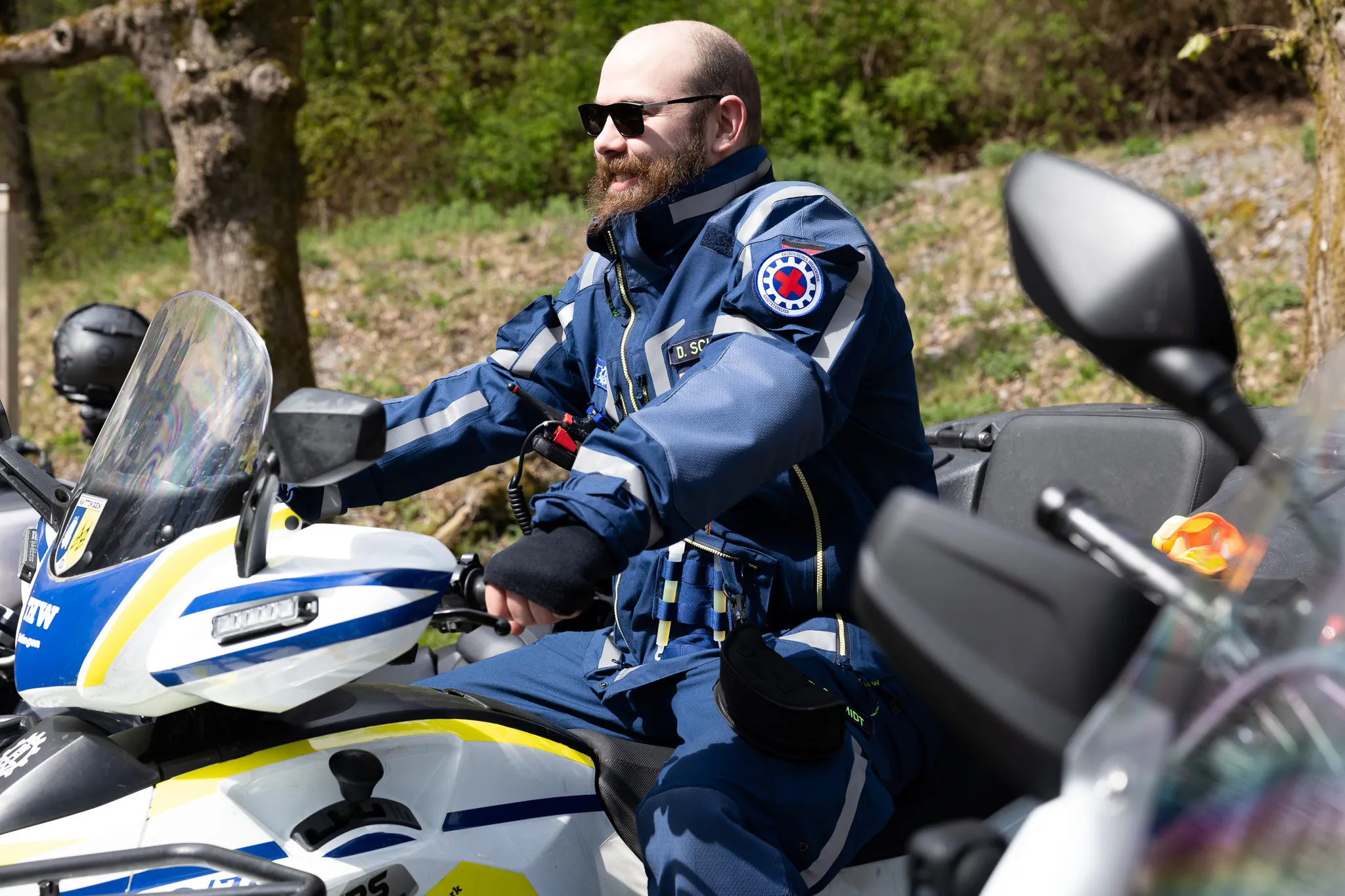 Ein Polizist mit Bart und Sonnenbrille sitzt auf einer gelben Motorrad, gekleidet in einer blauen Uniform mit Abzeichen und reflektierenden Streifen.