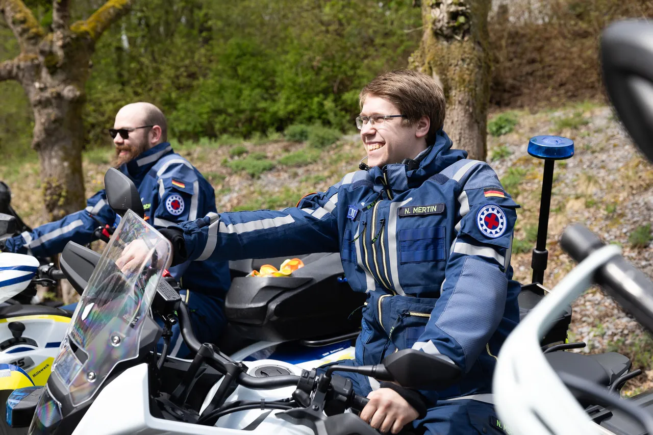 Zwei Polizisten in voller Motorradmontur sitzen auf ihren Motorrädern. Der vordere Polizist lächelt und deutet mit der Hand nach links.