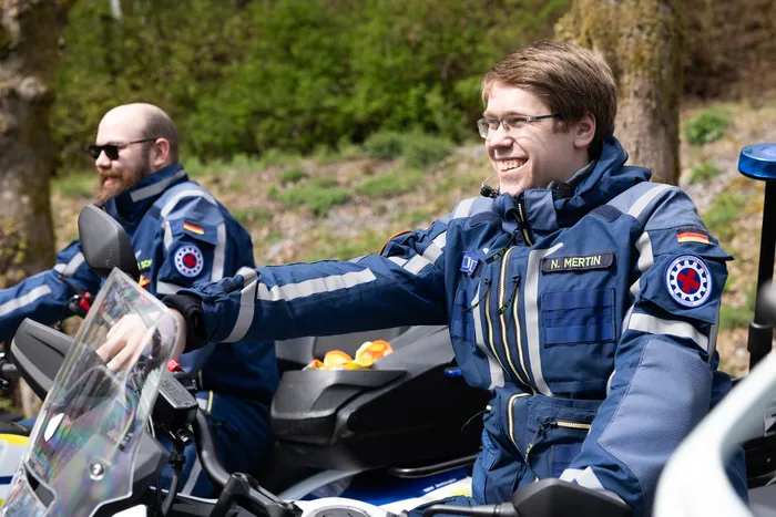 Zwei Polizisten in voller Motorradmontur sitzen auf ihren Maschinen und zeigen auf eine Karte. Im Hintergrund sind weitere Motorräder und Bäume zu sehen.