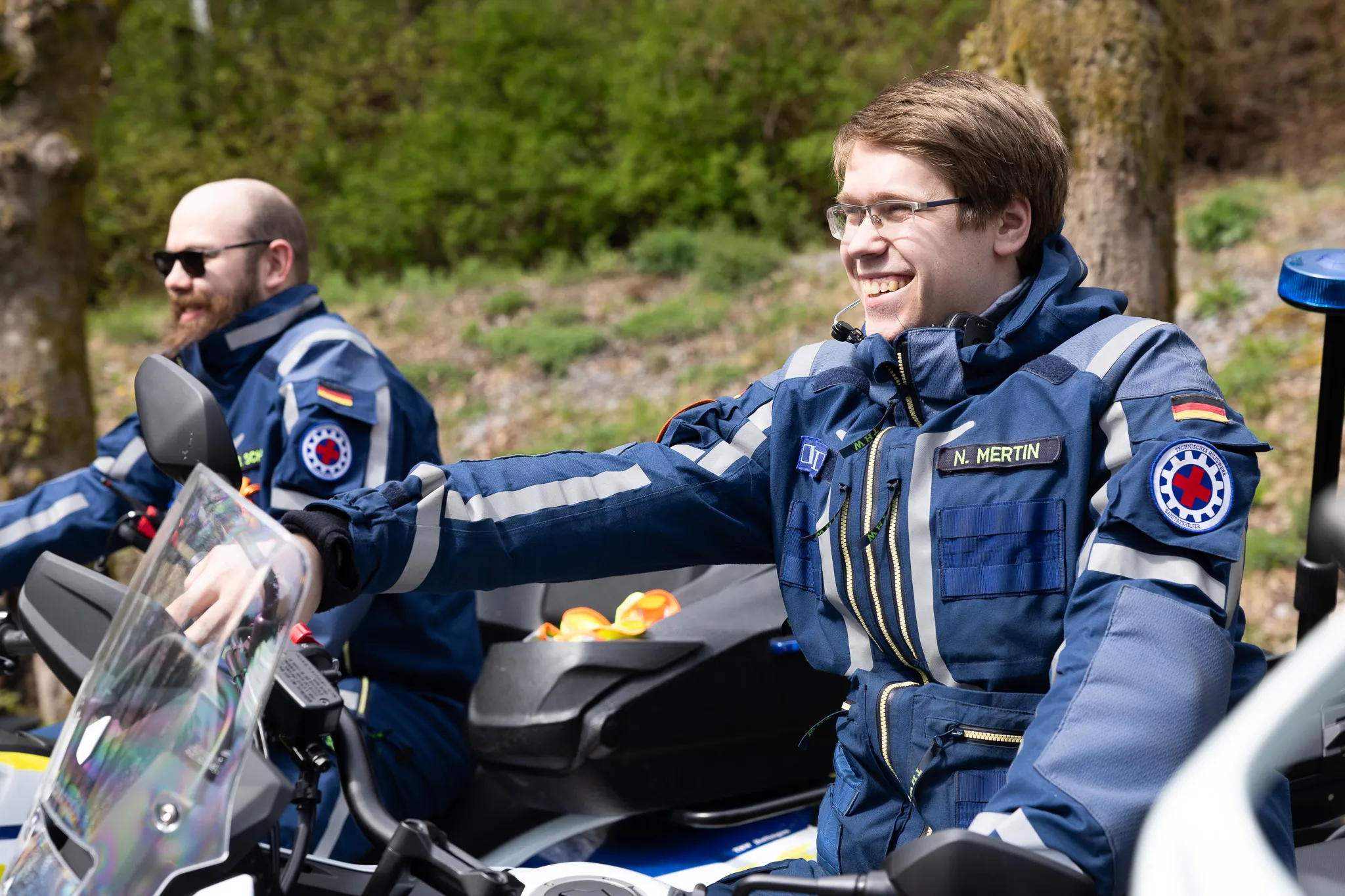 Zwei Polizisten in voller Motorradmontur sitzen auf ihren Maschinen und zeigen auf eine Karte. Im Hintergrund sind weitere Motorräder und Bäume zu sehen.