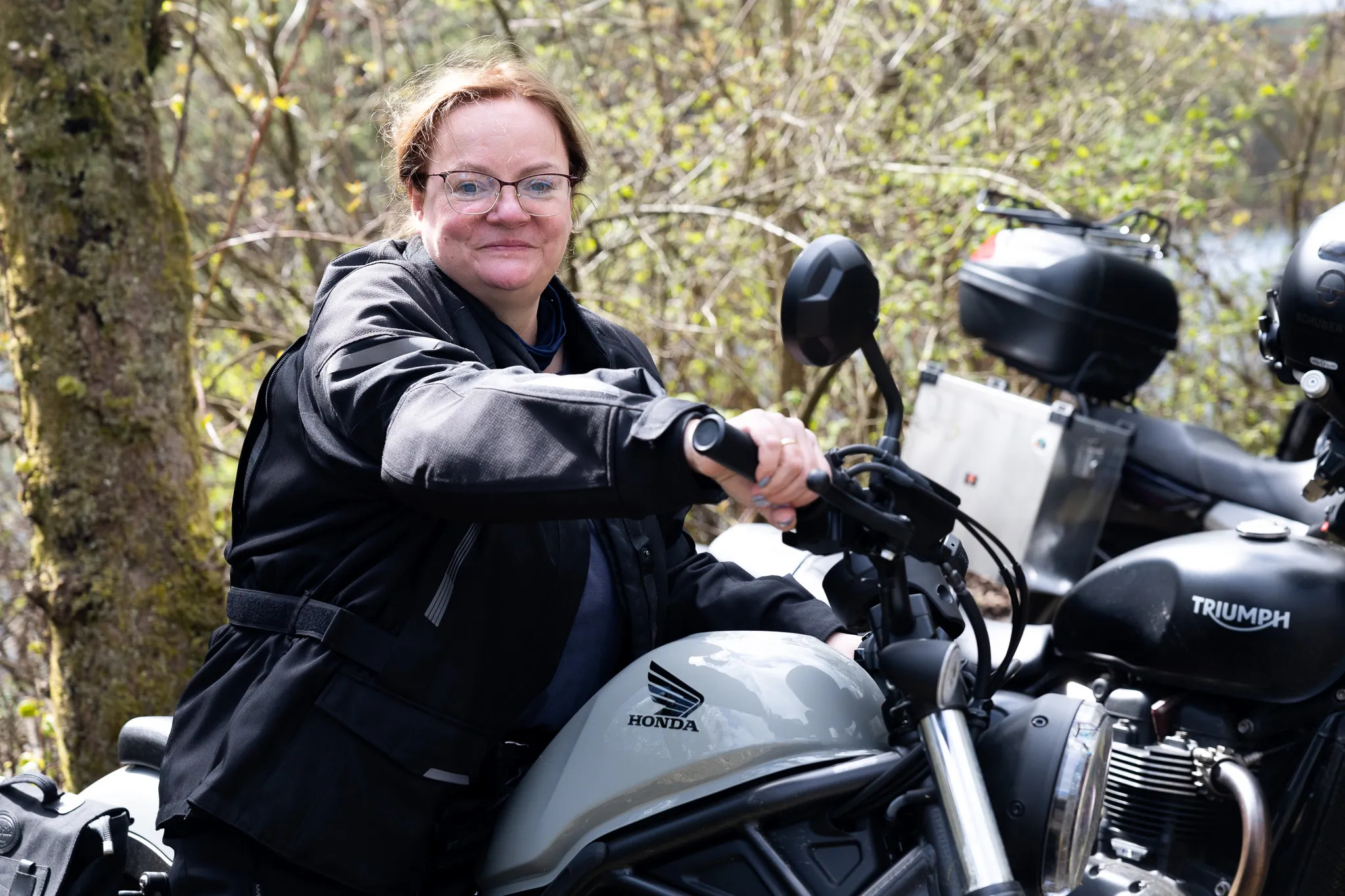 Eine Frau mit rotem Haar und Brille sitzt lächelnd auf einem Motorrad der Marke Honda. Im Hintergrund steht ein weiteres Motorrad der Marke Triumph und Bäume.