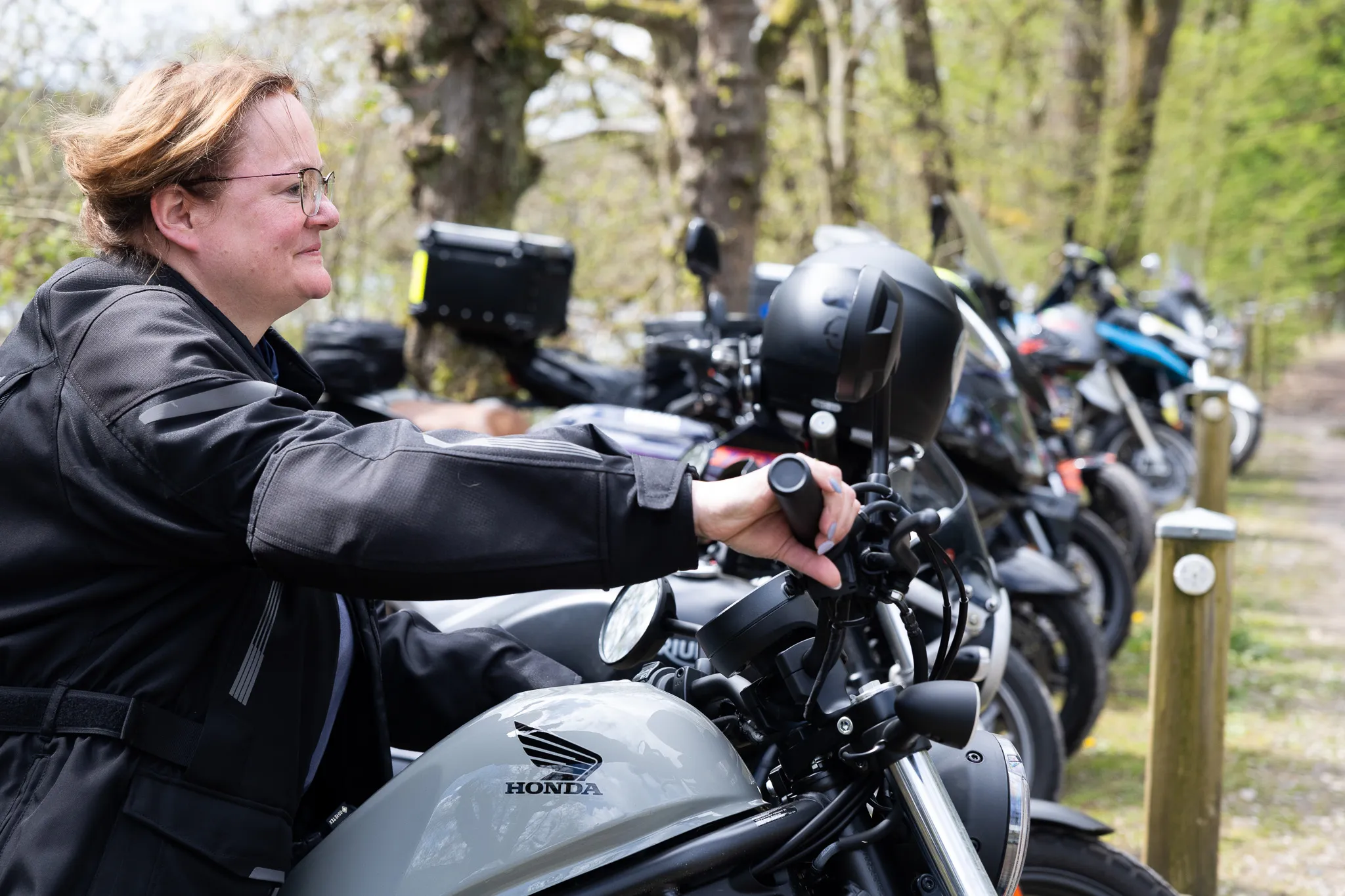 Eine Motorradfahrerin mit Brille und schwarzer Jacke sitzt auf einem silbernen Honda Motorrad. Im Hintergrund stehen weitere Motorräder und Bäume.