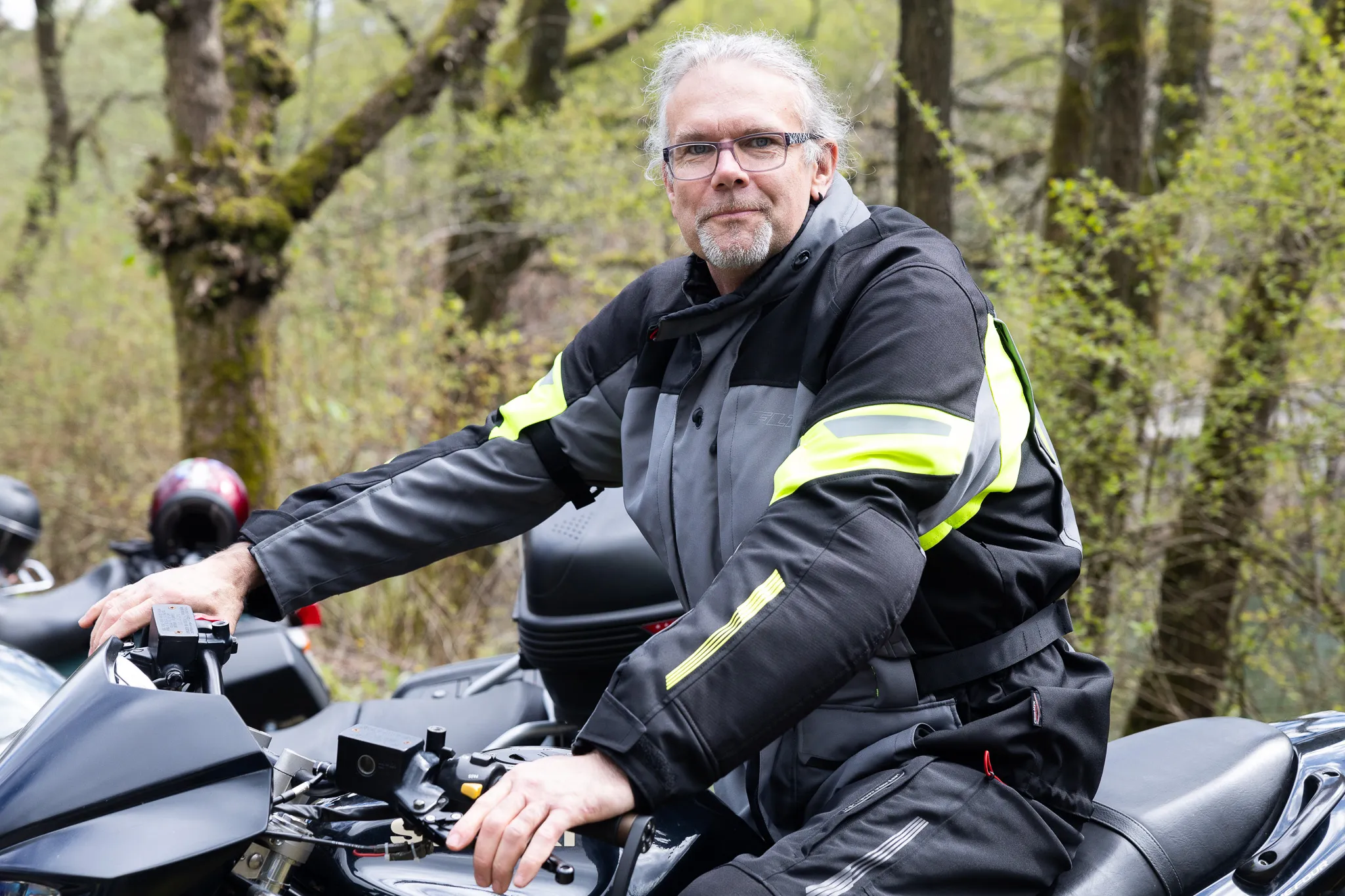 Mann mit grauen Haaren und Brille sitzt auf einem Motorrad und blickt in die Kamera. Er trägt Motorradbekleidung mit gelben Akzenten.