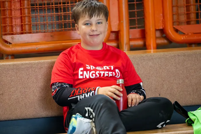 Ein lächelnder Junge in einem roten T-Shirt mit der Aufschrift „SPORT ENGSTEED“ sitzt auf einer Bank und hält eine rote Wasserflasche.