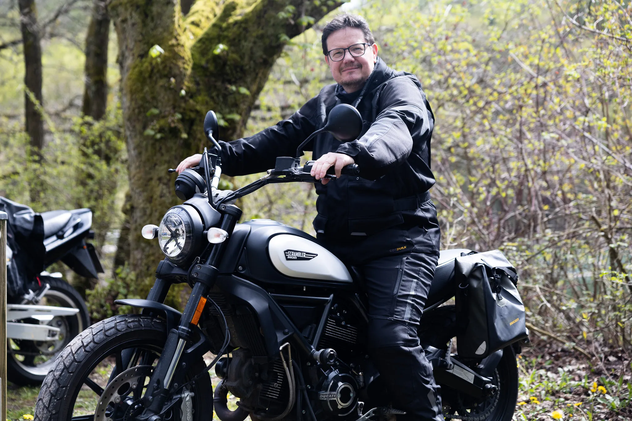 Ein Mann in Motorradkleidung sitzt auf einem schwarzen Motorrad und lächelt in die Kamera. Im Hintergrund sind weitere Motorräder und Bäume zu sehen.