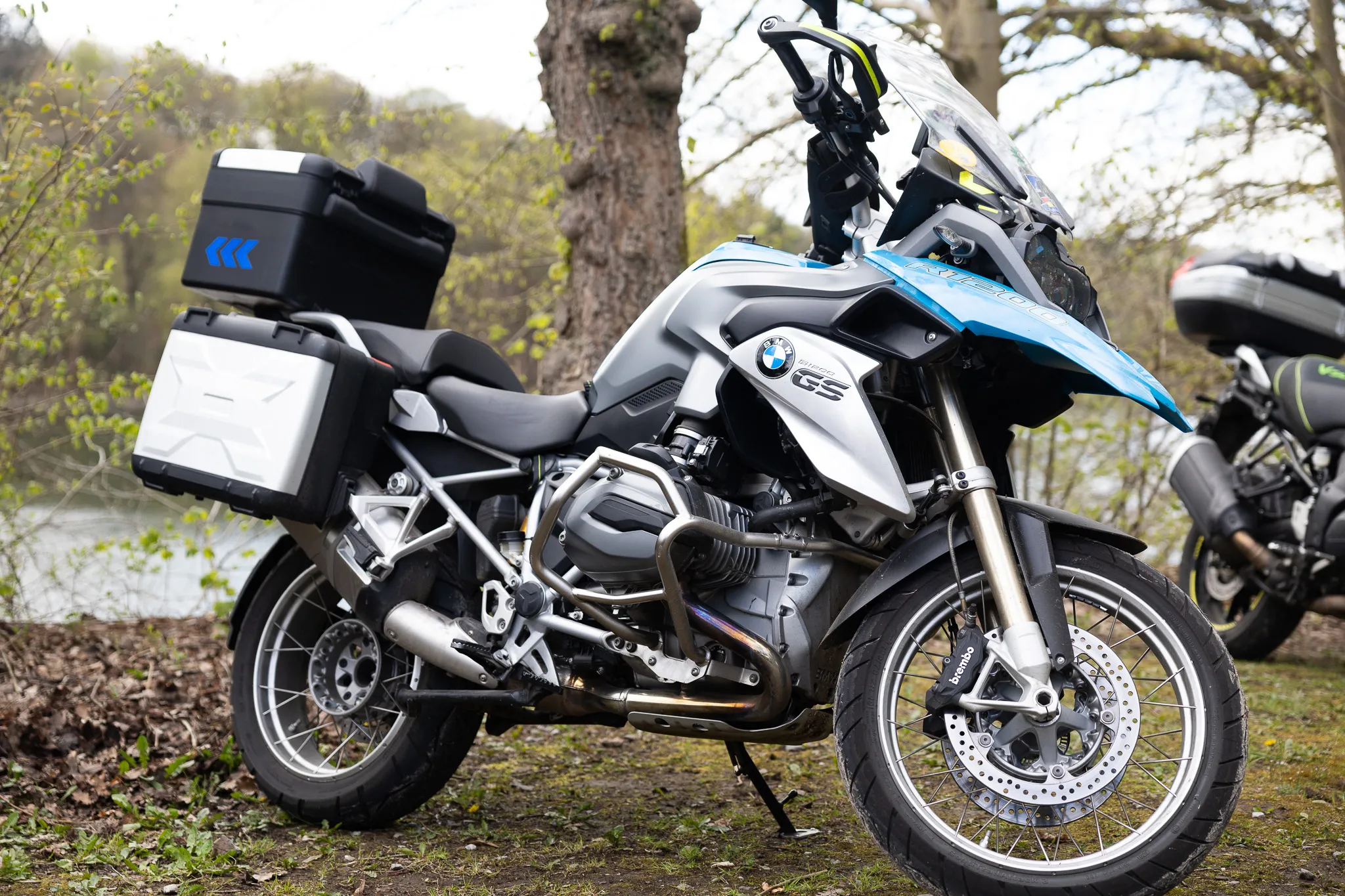 Silberne BMW-Motorrad mit Gepäckträgern steht an einem Ufer, im Hintergrund Bäume und ein weiteres Motorrad.