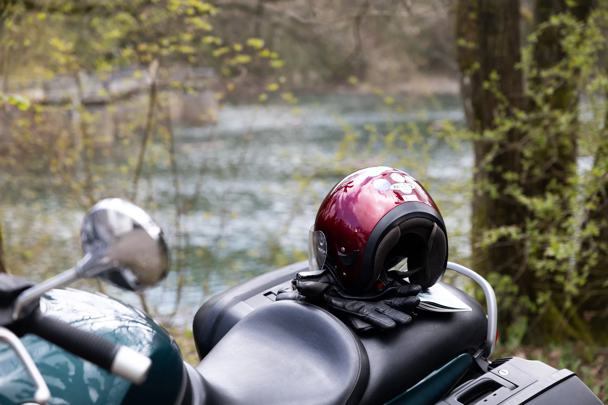 Roter Motorradhelm und schwarze Handschuhe liegen auf dem Gepäckträger eines grünen Motorrads vor einer Flusslandschaft.