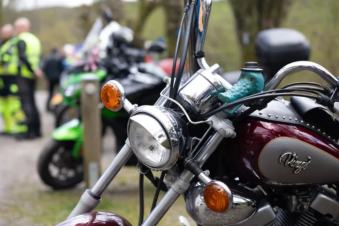 Nahaufnahme eines dunkelroten Motorrads mit Chromteilen und einem grünen Enten-Anhänger am Lenker. Im Hintergrund sind weitere Motorräder und Polizisten in Warnwesten zu sehen.
