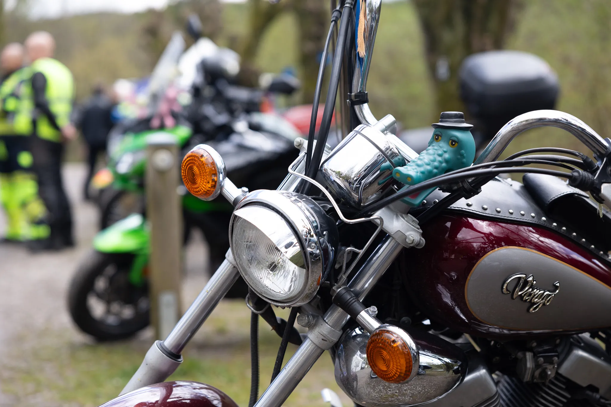 Nahaufnahme eines dunkelroten Motorrads mit Chromteilen und einem grünen Enten-Anhänger am Lenker. Im Hintergrund sind weitere Motorräder und Polizisten in Warnwesten zu sehen.