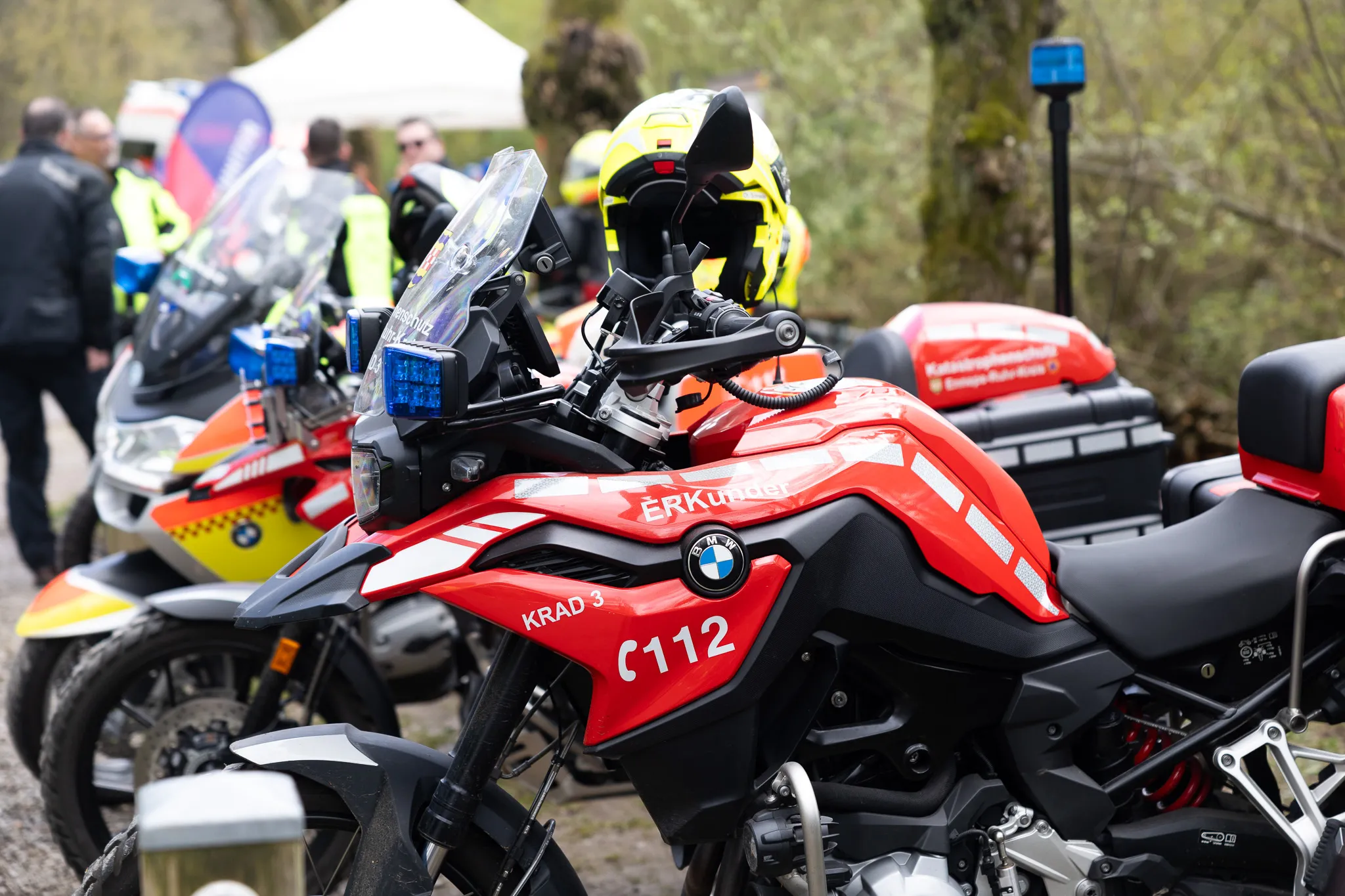 Rote BMW-Polizeimotorräder mit Blaulicht stehen auf einem Parkplatz, im Hintergrund sind weitere Einsatzkräfte und ein Zelt zu sehen.
