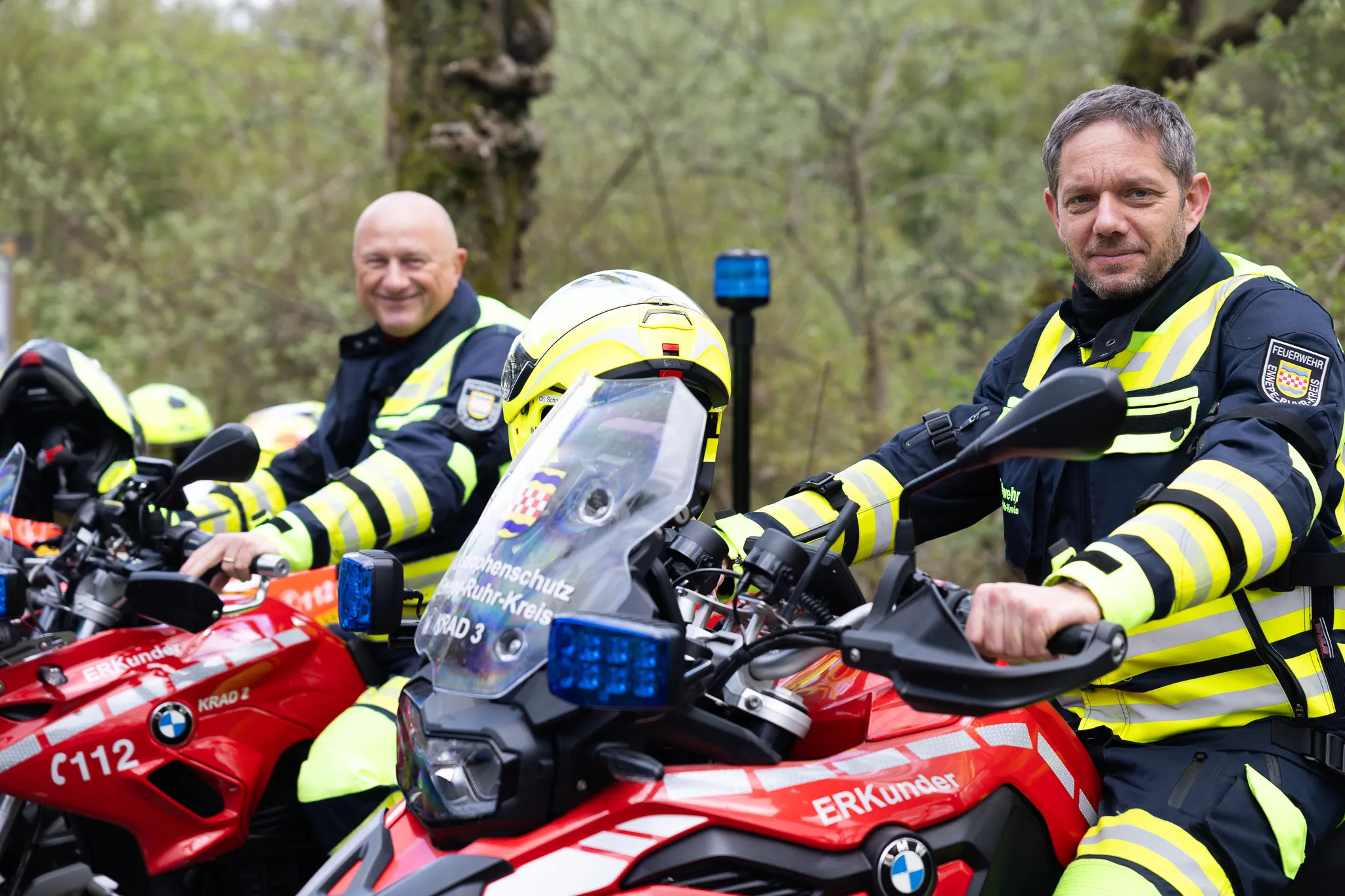 Zwei Feuerwehrleute in voller Montur sitzen auf roten BMW-Motorrädern mit Blaulicht und Aufschrift „Erkunder“ und „Feuerwehr“. Im Hintergrund ein weiterer Motorradfahrer.