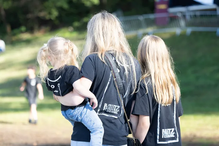 Eine Frau trägt ein kleines Kind auf dem Arm, neben ihr steht ein Mädchen. Alle tragen schwarze T-Shirts mit dem Aufdruck „BAUTZ FESTIVAL“.