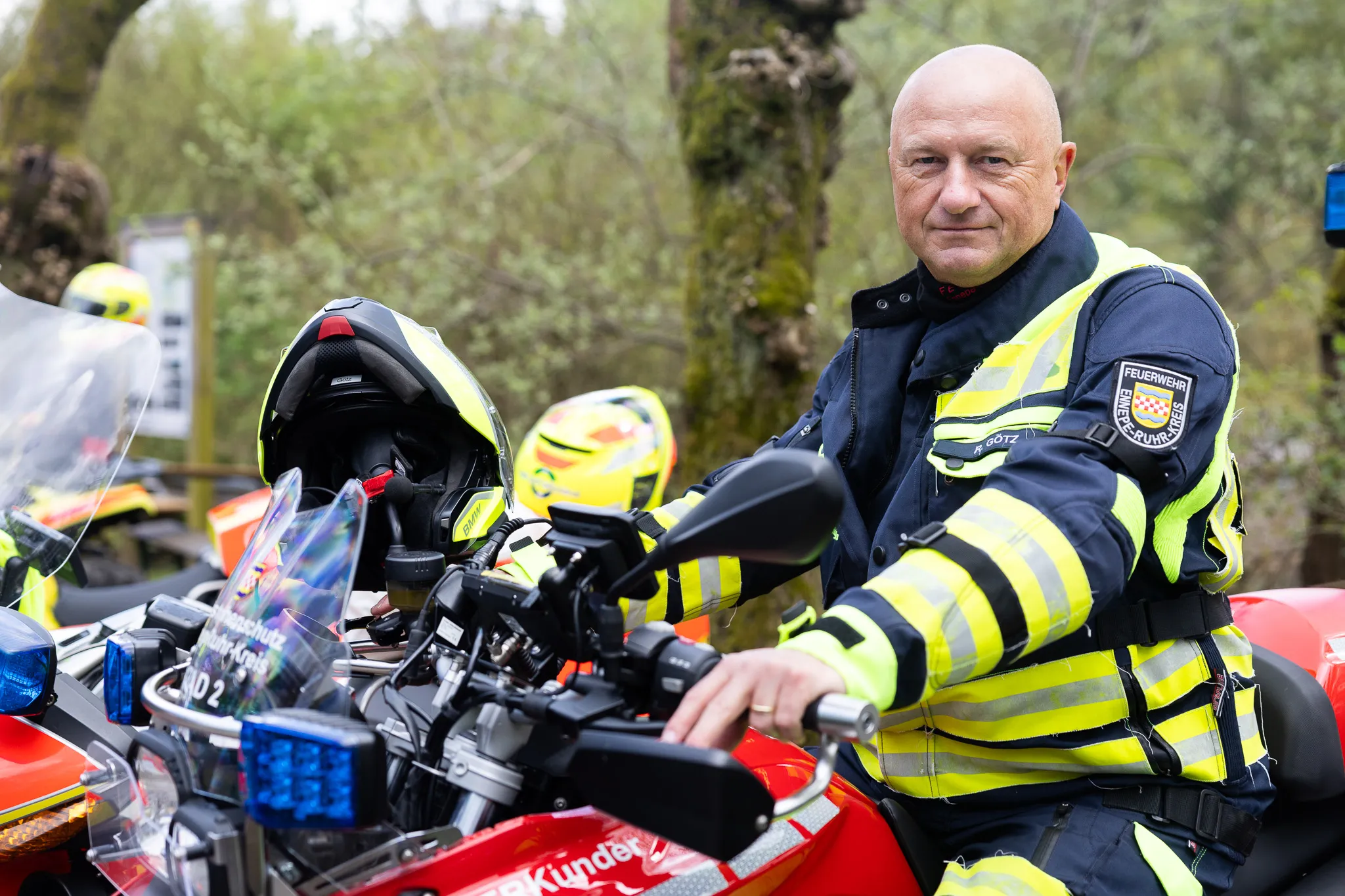 Feuerwehrmann in Einsatzuniform sitzt auf einem roten Motorrad mit Blaulicht und Helm. Im Hintergrund sind weitere Einsatzfahrzeuge und Bäume zu sehen.