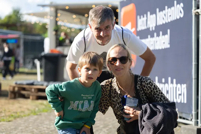 Ein Mann, eine Frau mit Sonnenbrille und ein kleiner Junge in einem grünen Sweatshirt posieren gemeinsam vor einem Banner im Freien.