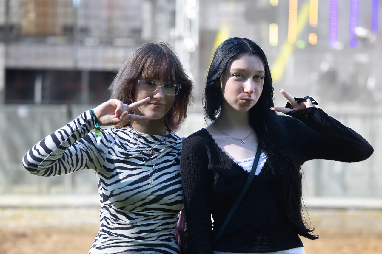 Zwei junge Frauen posieren mit Peace-Zeichen; eine trägt eine Brille und ein Zebra-Muster-Shirt, die andere ein schwarzes Oberteil.