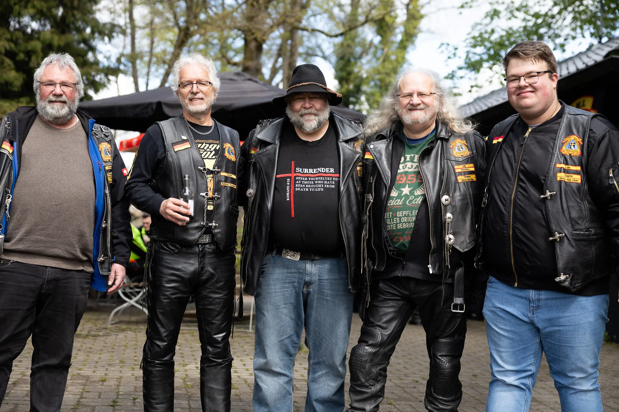 Fünf Männer mittleren Alters mit Biker-Kleidung und -Abzeichen stehen lächelnd auf einem Platz. Im Hintergrund sind weitere Personen und Zelte zu sehen.
