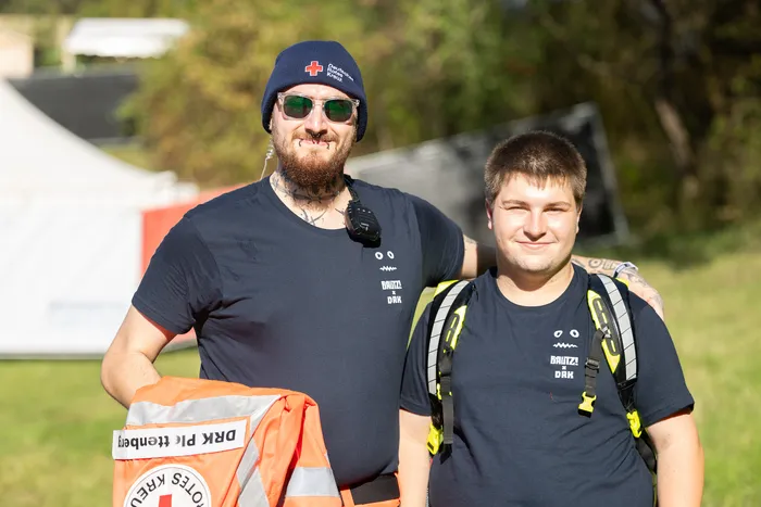 Vincent Sonore mit blauem Beanie und Sonnenbrille hält eine orangefarbene DRK-Tasche, neben ihm steht JP Lekic.