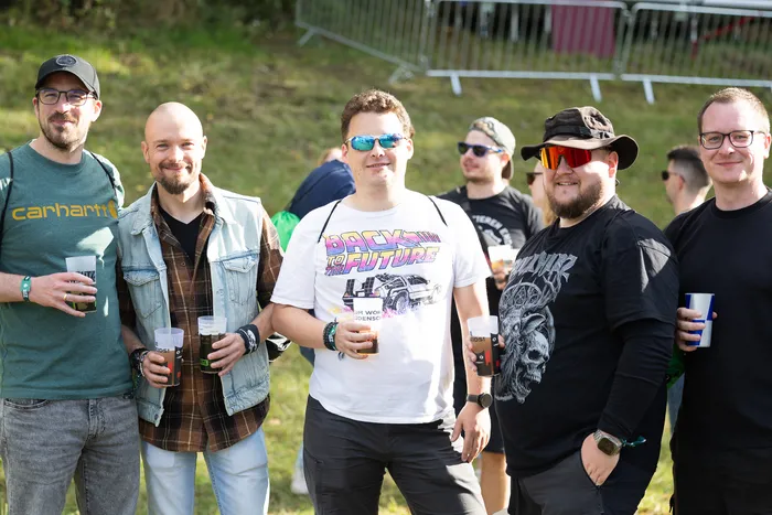 Sechs Männer stehen nebeneinander im Freien und halten Getränke in Bechern. Einer trägt ein weißes T-Shirt mit einem „Back to the Future“-Motiv.