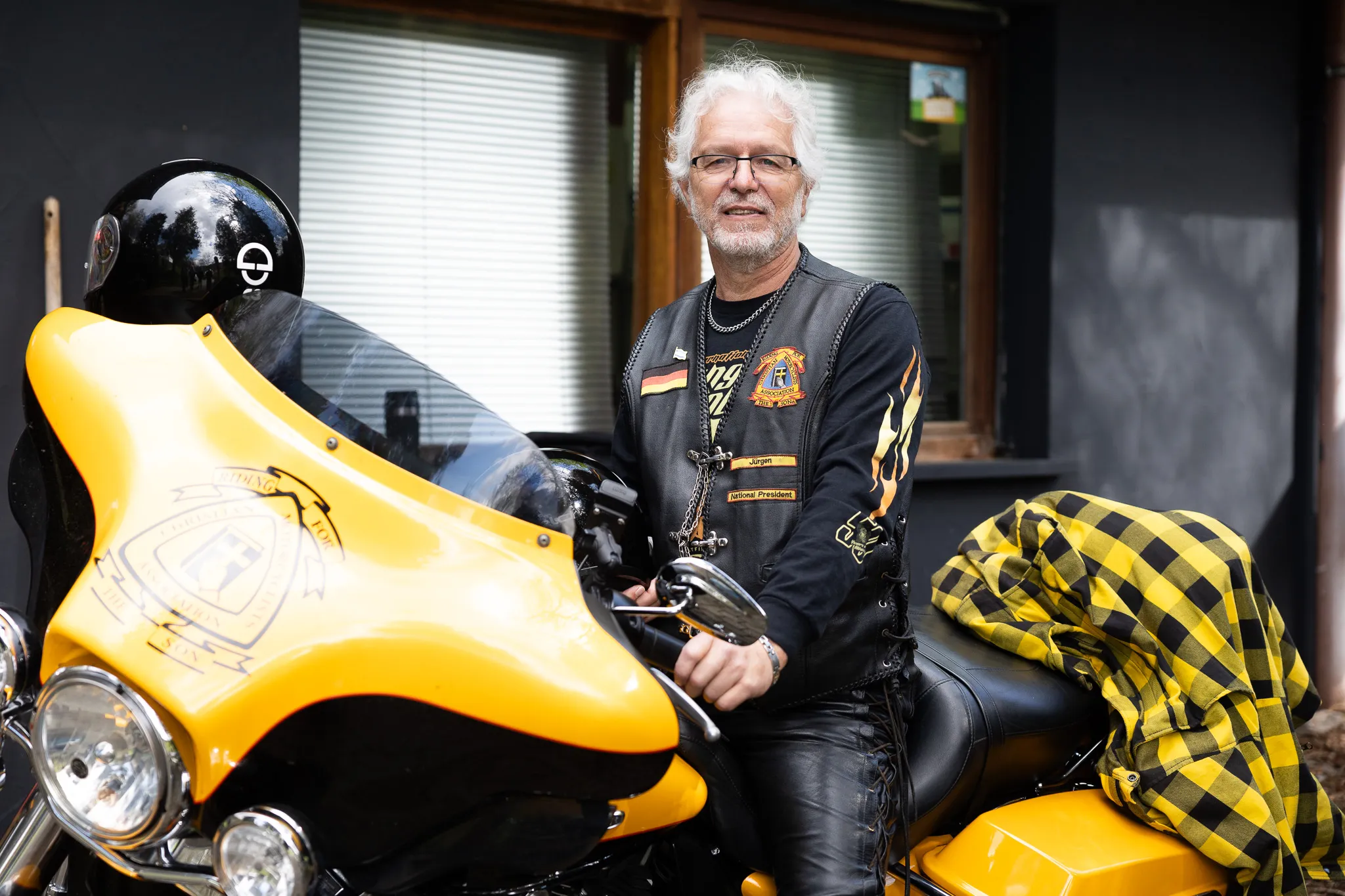 Ein Mann mit grauen Haaren und Bart sitzt auf einem gelben Motorrad vor einem Haus. Er trägt eine Lederweste mit Aufnähern und eine Sonnenbrille.