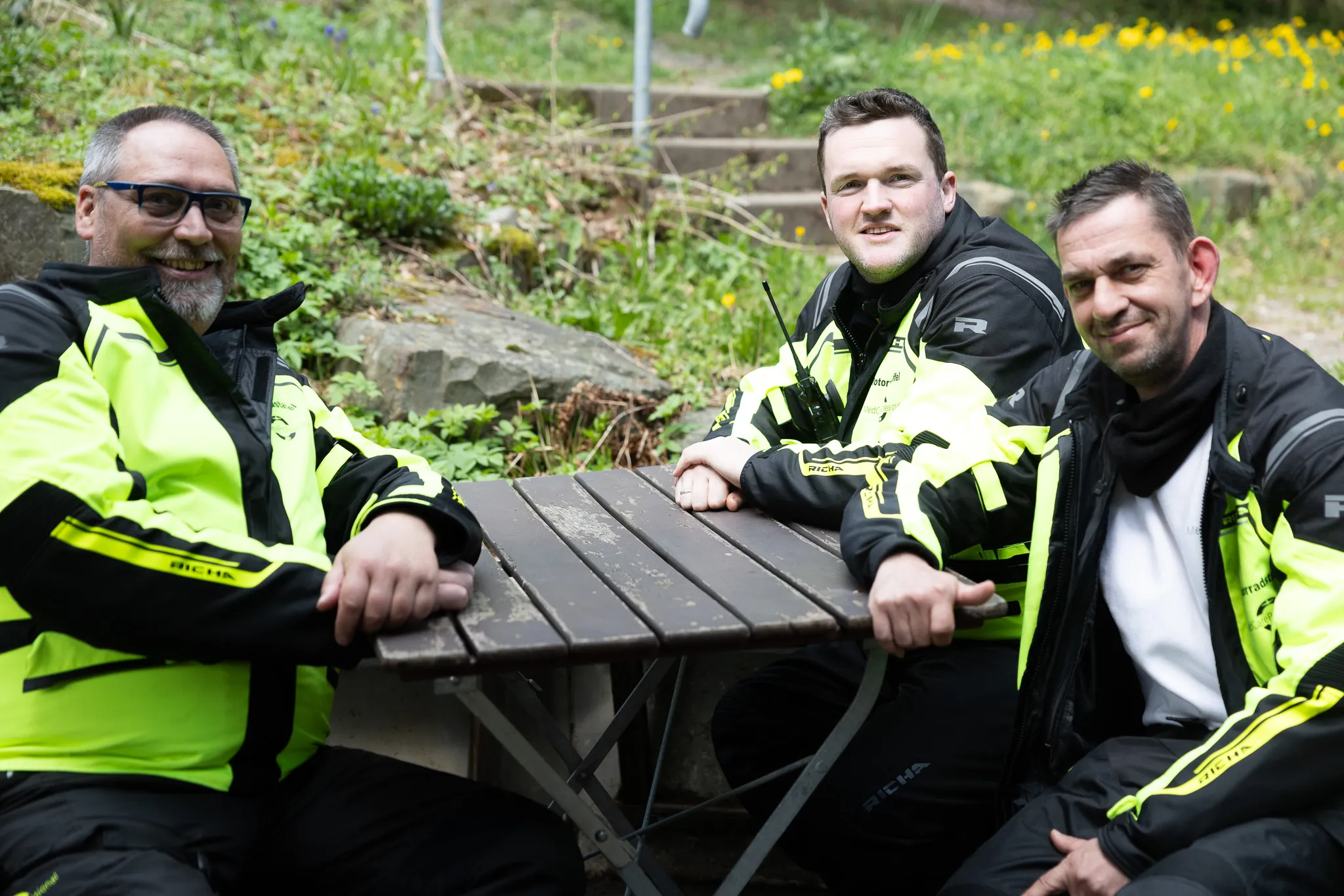 Drei Polizisten in fluoreszierender Schutzkleidung sitzen an einem Picknicktisch im Freien und lächeln in die Kamera. Im Hintergrund blühende Pflanzen und Büsche.