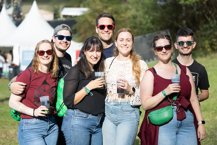 Sechs lächelnde Personen mit Sonnenbrillen posieren bei einem Outdoor-Event und halten Getränke in Plastikbechern.