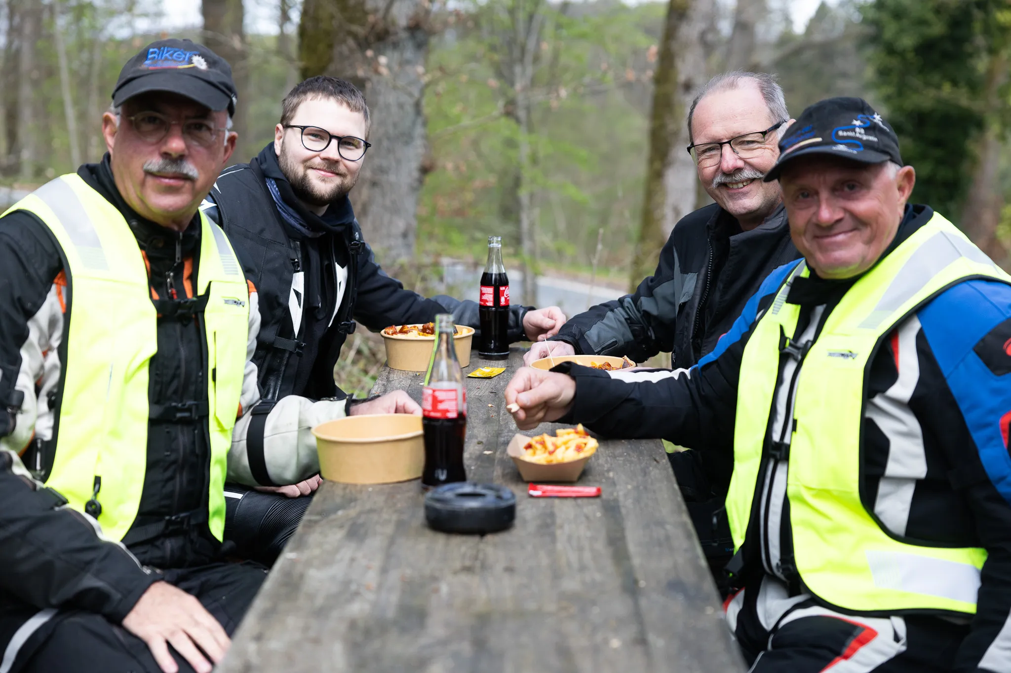 Vier Motorradfahrer sitzen an einem Holztisch und essen Pommes aus Schalen, Getränke stehen bereit. Sie tragen Motorradkleidung und Warnwesten.