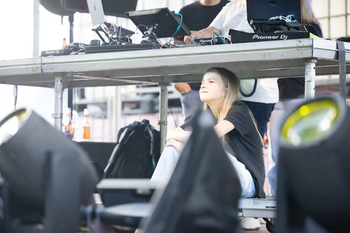 Eine junge Frau sitzt auf einer Bühne und blickt nach oben, während im Vorder- und Hintergrund DJ-Equipment und Bühnenlichter zu sehen sind.