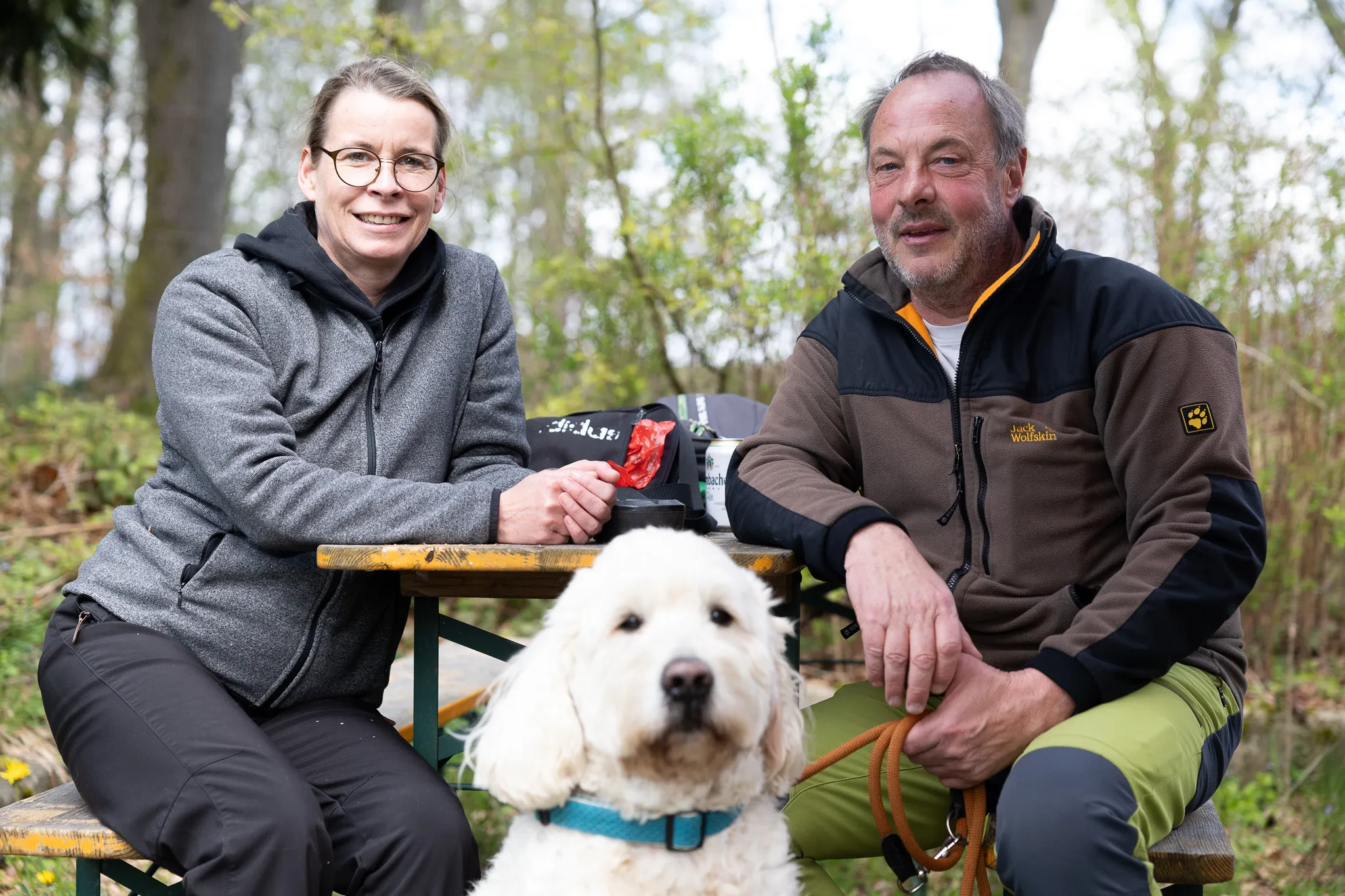 Zwei Personen sitzen an einem Picknicktisch im Freien, ein mittelgroßer, lockiger Hund blickt in die Kamera. Im Hintergrund Bäume und Büsche.