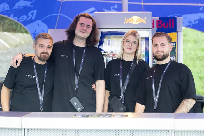 Vier Personen in schwarzen T-Shirts mit „Rotwer Events“-Aufdruck und Lanyards stehen hinter einem Tresen vor einem Red Bull Kühlschrank.