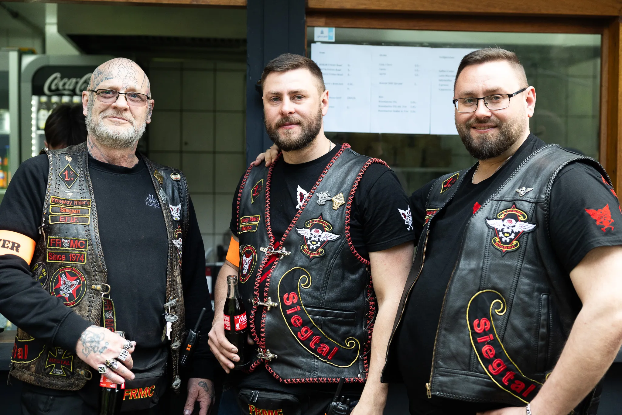Drei Männer mit Biker-Kutten und Patches stehen lächelnd vor einer Schaufensterfront. Einer hält eine Flasche in der Hand.
