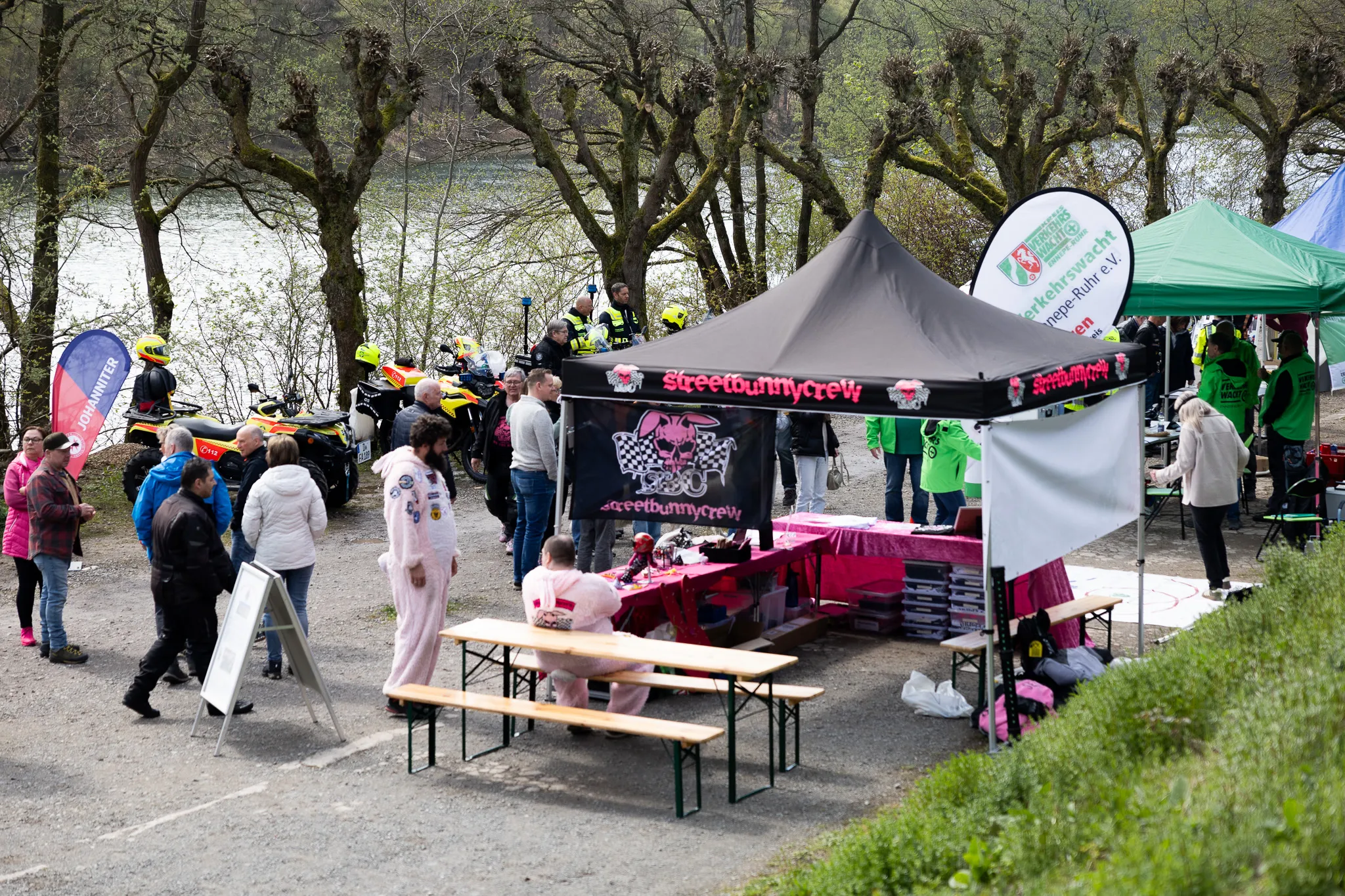 Ein Stand mit dem Aufdruck „Streetbunnycrew“ steht an einem Ufer, umgeben von Menschen und Motorrädern. Im Hintergrund sind Polizisten in Warnwesten zu sehen.