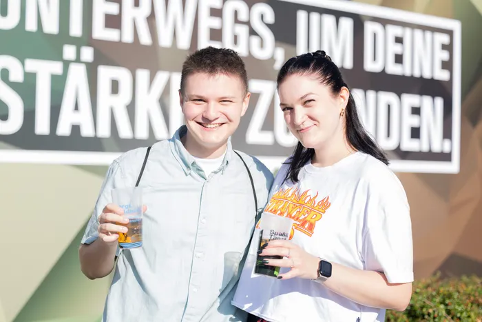 Vincent Sonore und JP Lekic lächeln vor einem Schild mit der Aufschrift „UNTERWEGS, UM DEINE STÄRKE ZÜNDEN.“.