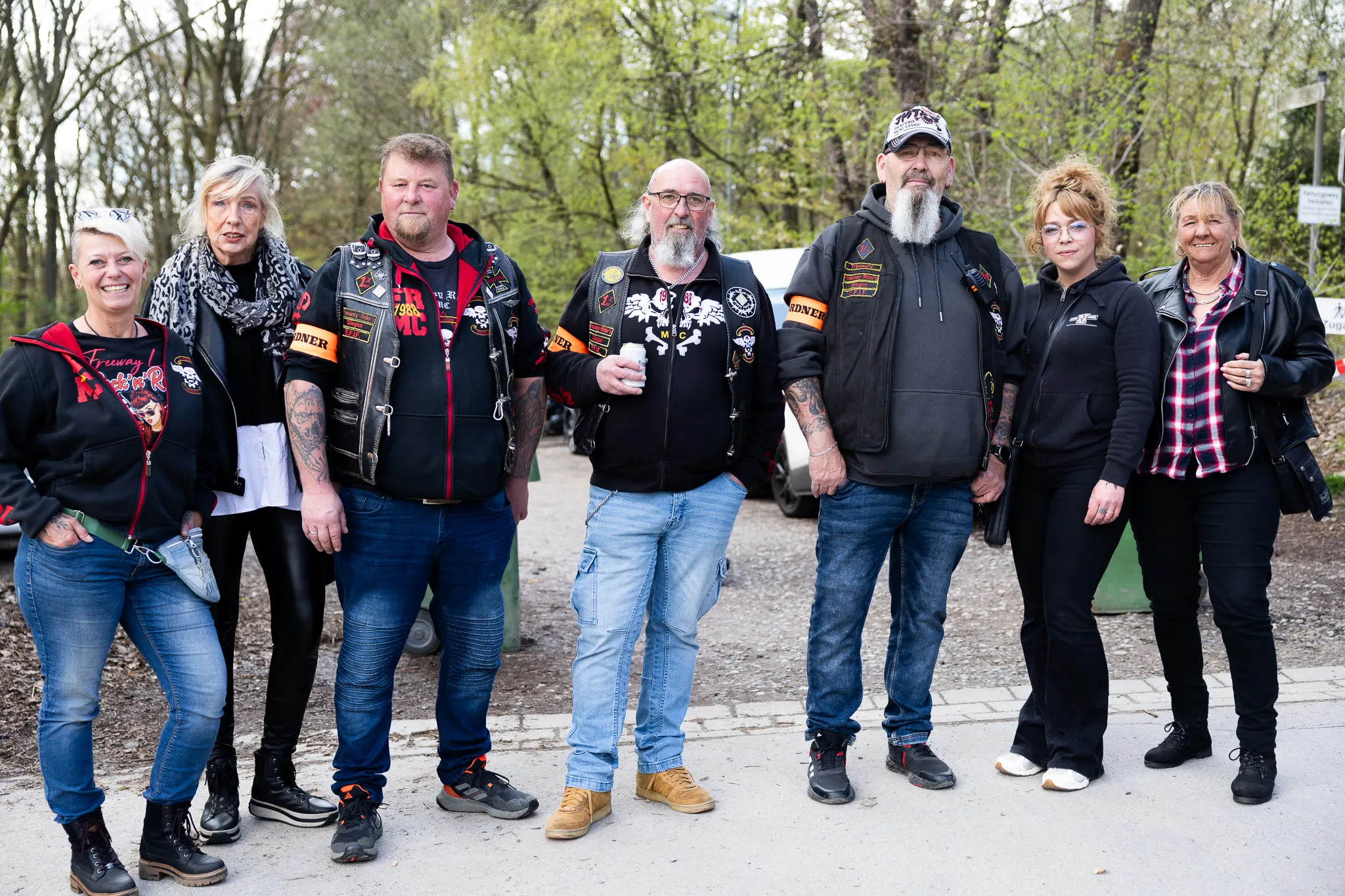 Sieben Personen, darunter mehrere Motorradfahrer mit Lederjacken und Aufnähern, stehen vor Motorrädern und posieren für ein Foto im Freien.