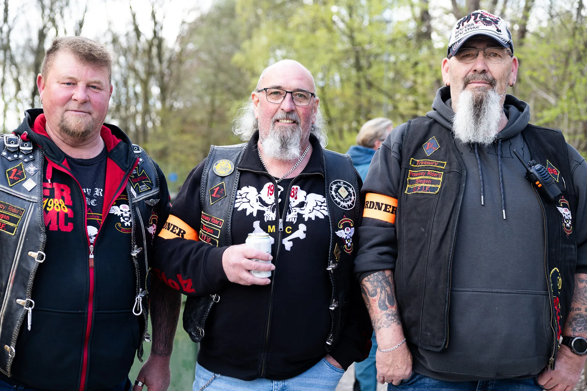 Drei Männer mit langen Bärten und Lederwesten mit Aufnähern stehen nebeneinander. Im Hintergrund verschwommene Menschen und Bäume.