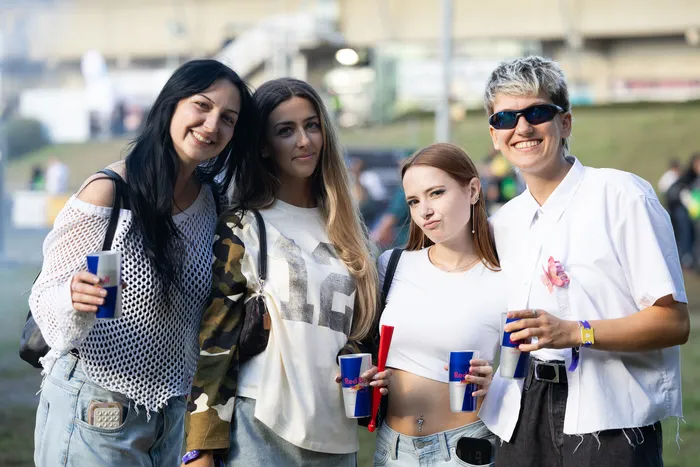 Vier junge Frauen lächeln in die Kamera und halten Getränkebecher bei einem Open-Air-Event.