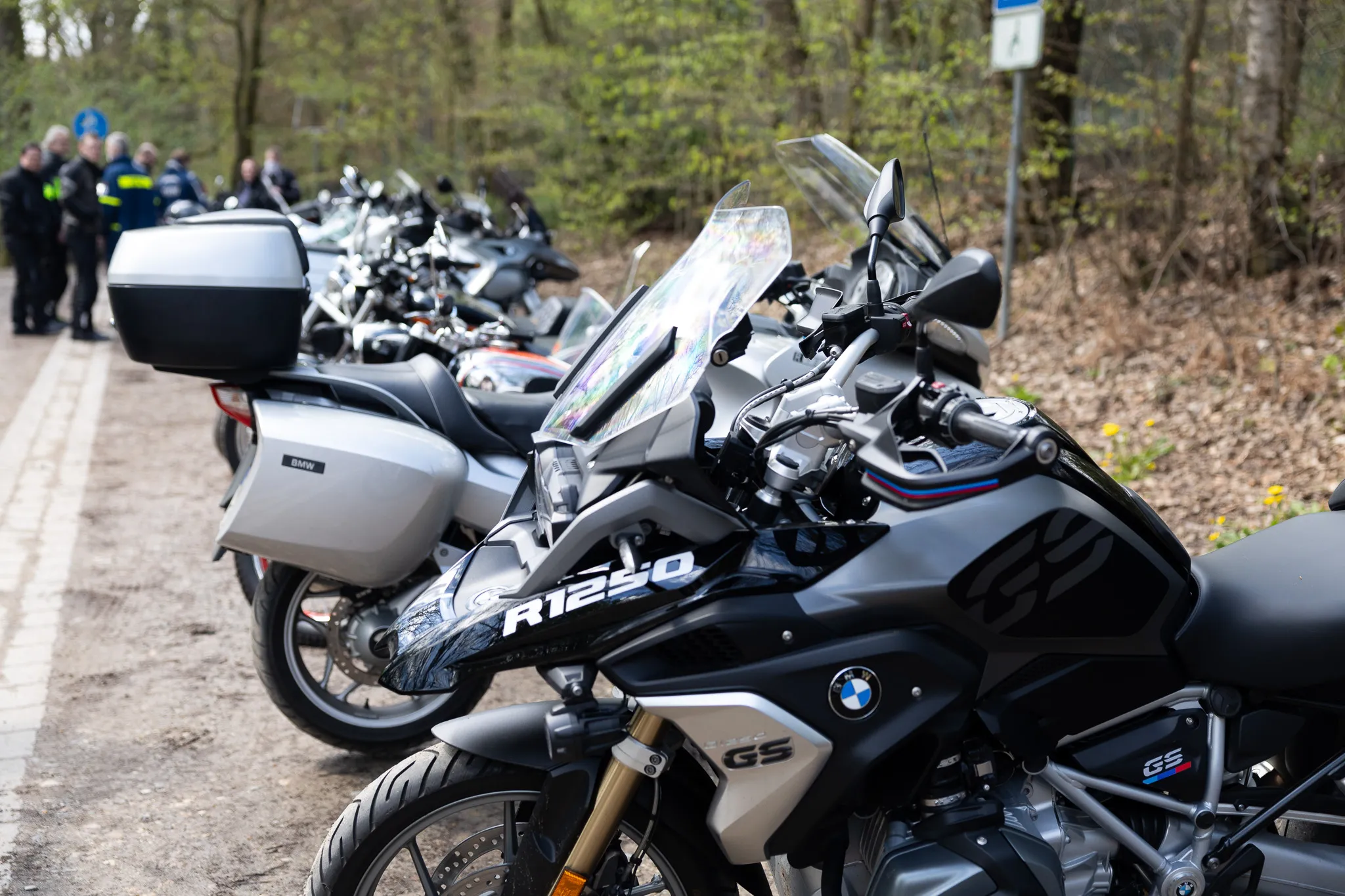 Mehrere schwarze BMW Motorräder stehen auf einem Kiesweg, im Hintergrund Polizisten und Bäume.