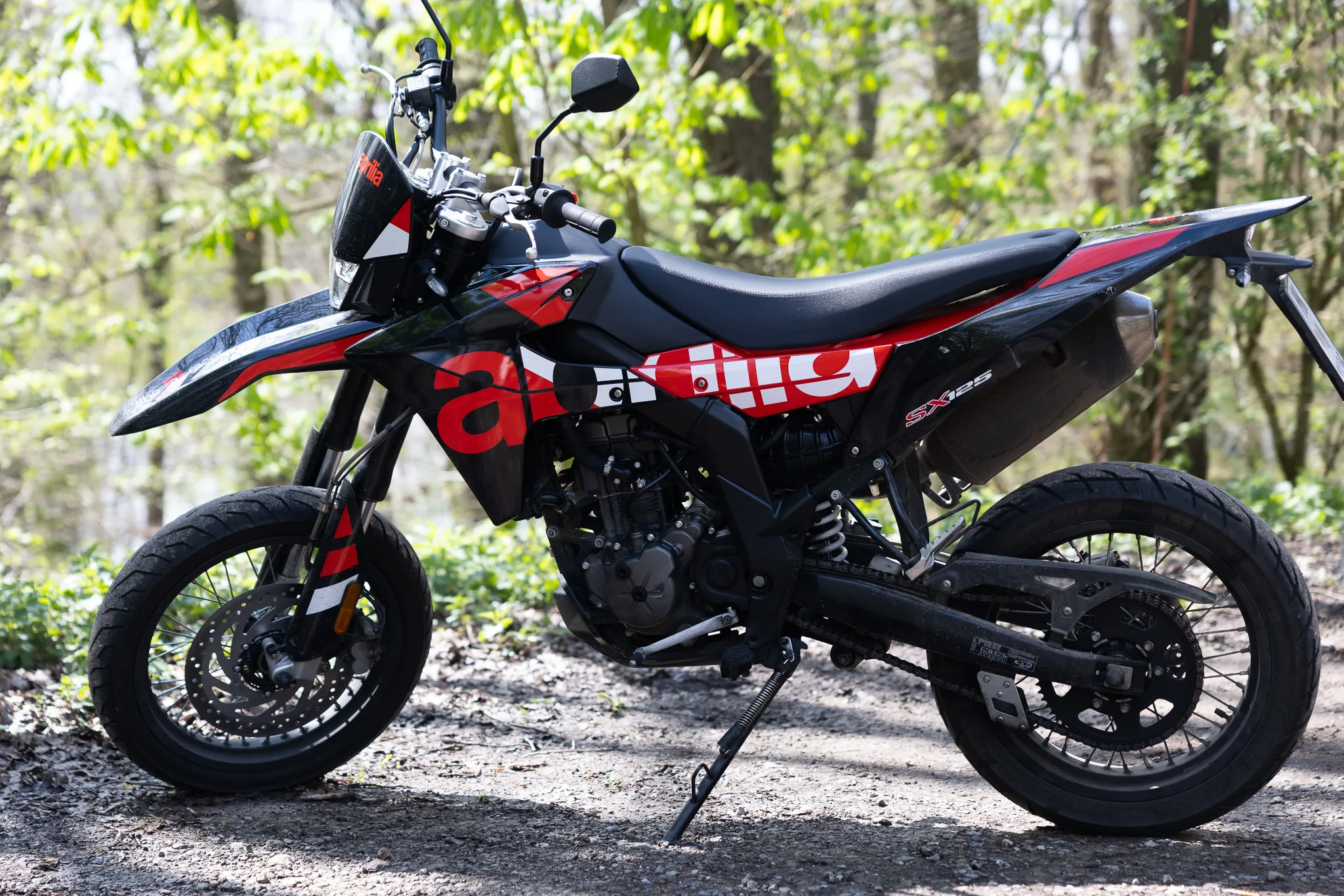 Schwarze und rote Aprilia SX 125 Motorrad steht auf einem Schotterweg vor einem Wald. Das Motorrad trägt das Logo der italienischen Firma Aprilia und das Kürzel SX 125.