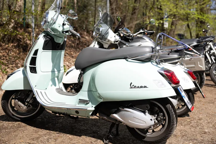 Hellgrüner Vespa Roller steht neben anderen Motorrädern auf einem Kiesparkplatz, im Hintergrund Bäume.
