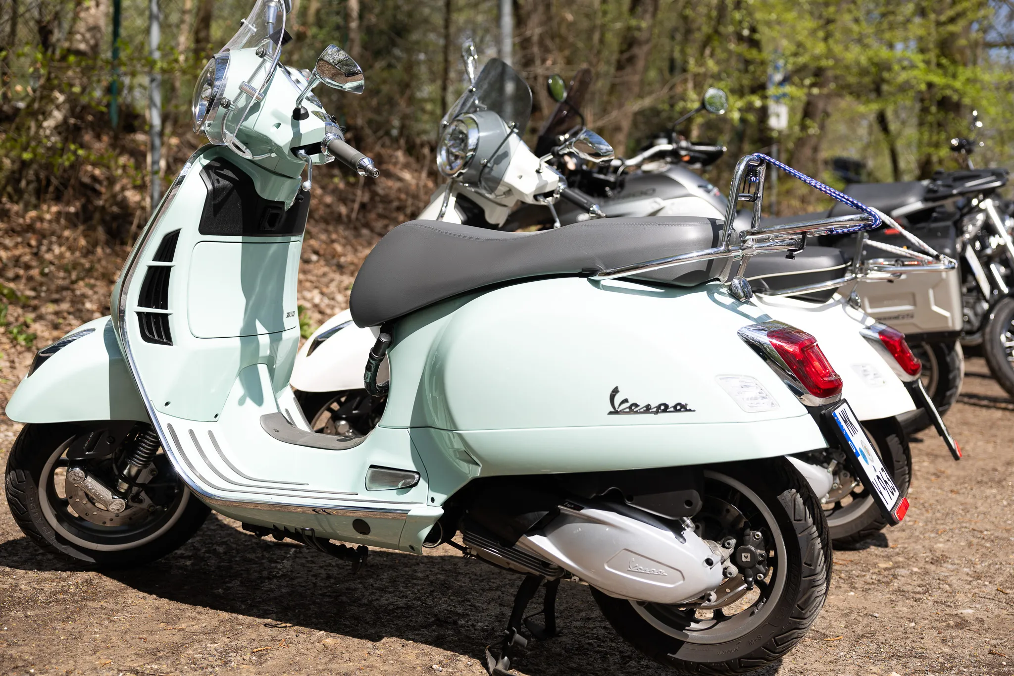 Hellgrüner Vespa Roller steht neben anderen Motorrädern auf einem Kiesparkplatz, im Hintergrund Bäume.