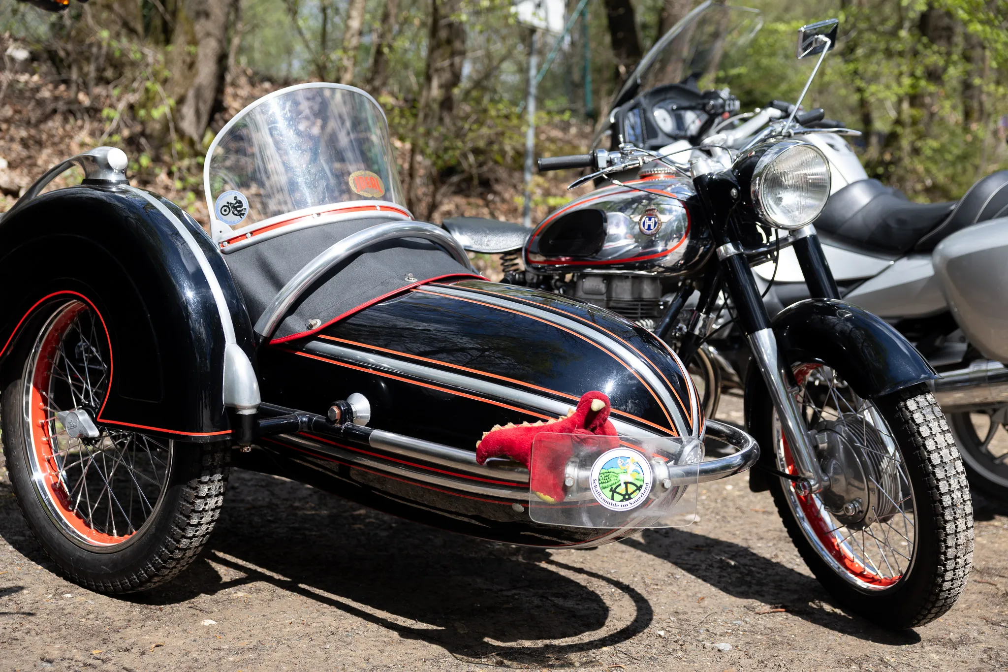 Motorrad mit Beiwagen, verziert mit roten Socken und einem Aufkleber, steht auf einem Parkplatz vor Bäumen. Im Hintergrund sind weitere Motorräder zu sehen.