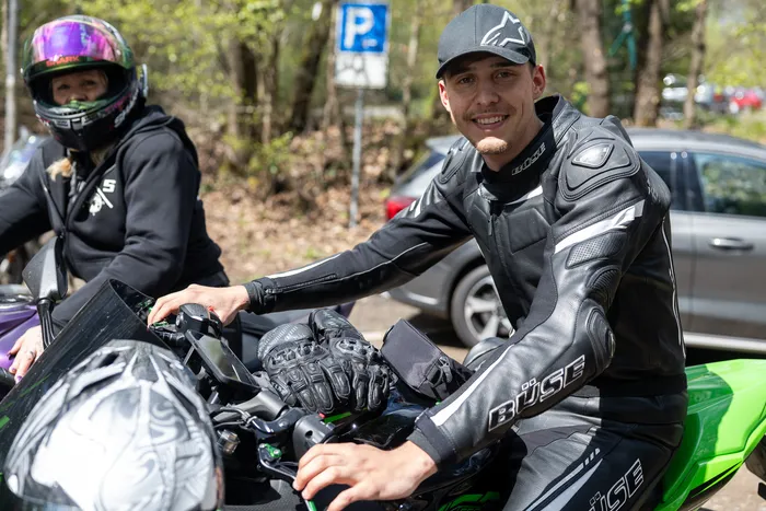 Motorradfahrer in Schutzkleidung sitzt lächelnd auf seinem Motorrad, im Hintergrund eine weitere Person auf einem Motorrad und ein Parkschild.