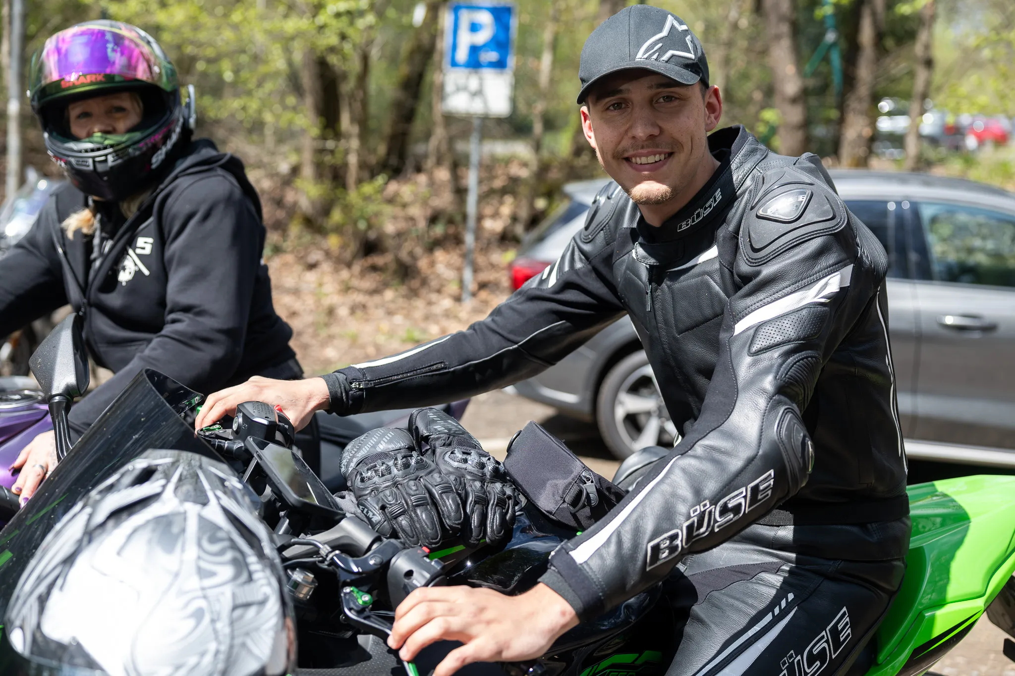 Motorradfahrer in Schutzkleidung sitzt lächelnd auf seinem Motorrad, im Hintergrund eine weitere Person auf einem Motorrad und ein Parkschild.