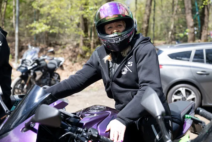 Eine Motorradfahrerin mit Helm sitzt auf ihrem Motorrad. Im Hintergrund sind weitere Motorräder und ein grauer PKW zu sehen.