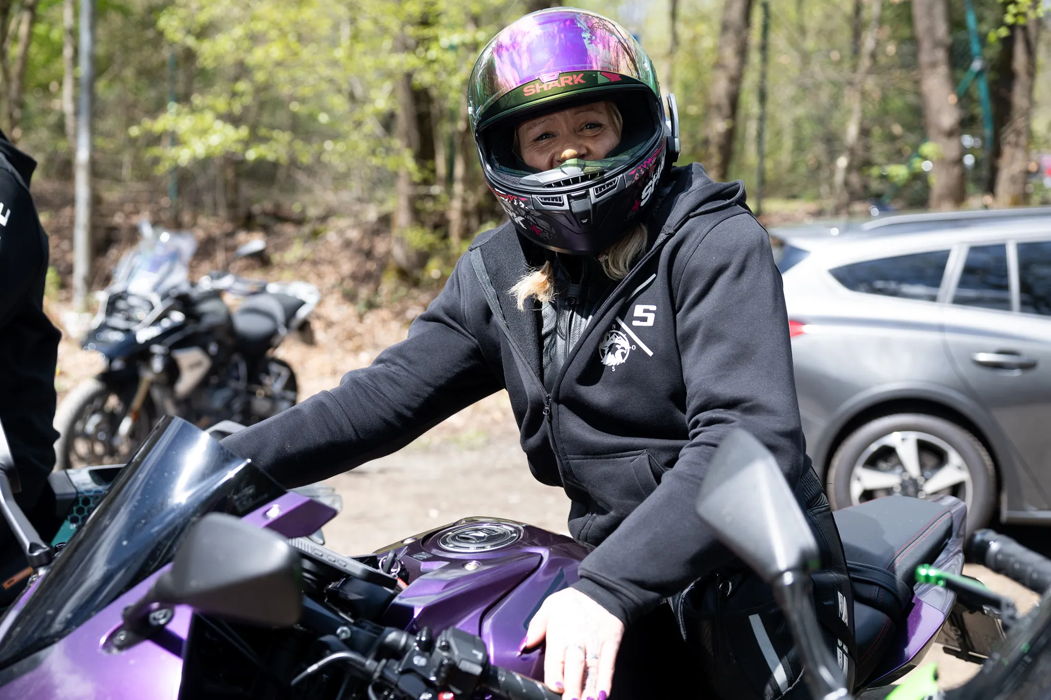 Eine Motorradfahrerin mit Helm sitzt auf ihrem Motorrad. Im Hintergrund sind weitere Motorräder und ein grauer PKW zu sehen.