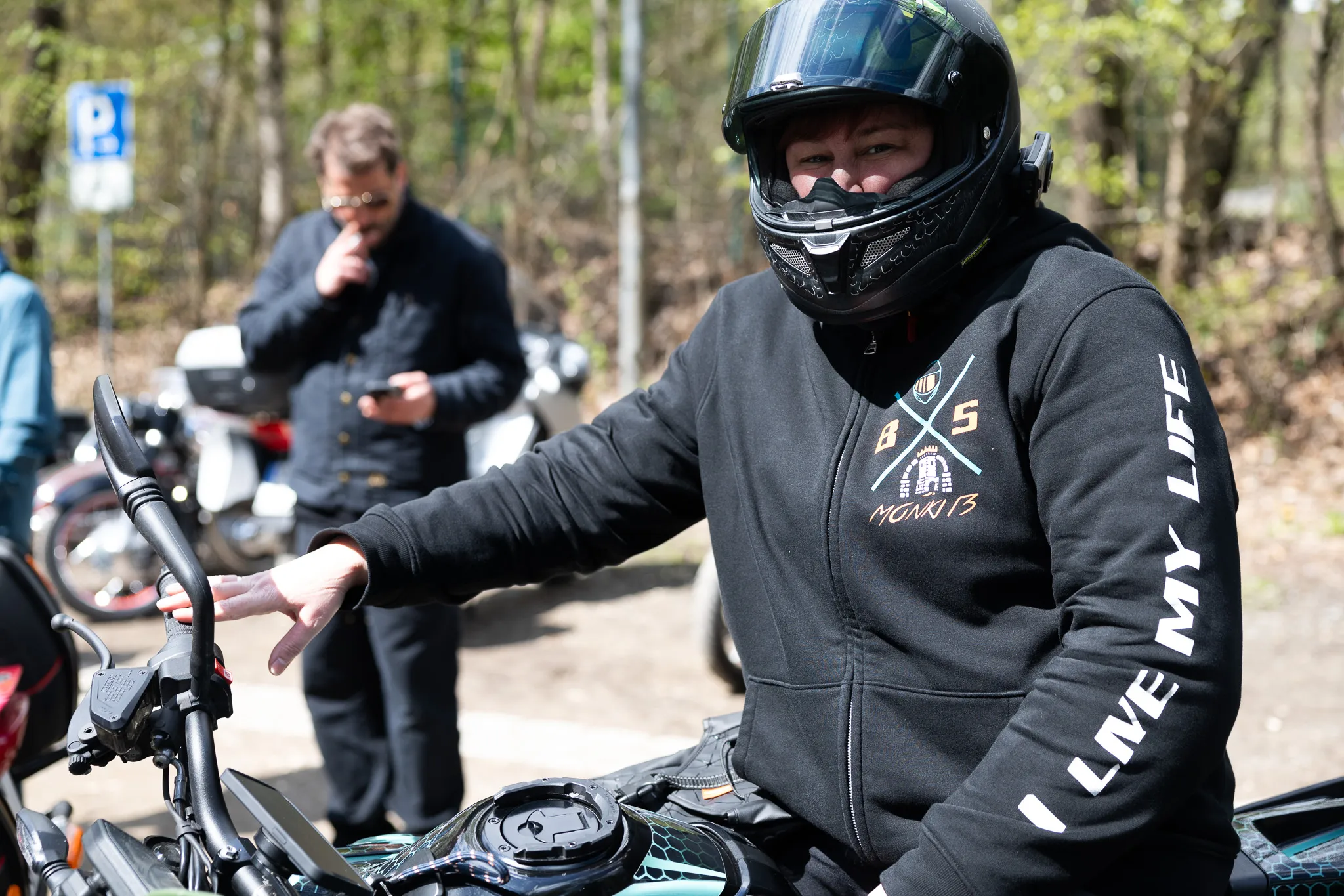 Motorradfahrer mit Helm und schwarzer Jacke zeigt mit der Hand auf etwas vor ihm, im Hintergrund ein weiterer Mann und Motorräder.