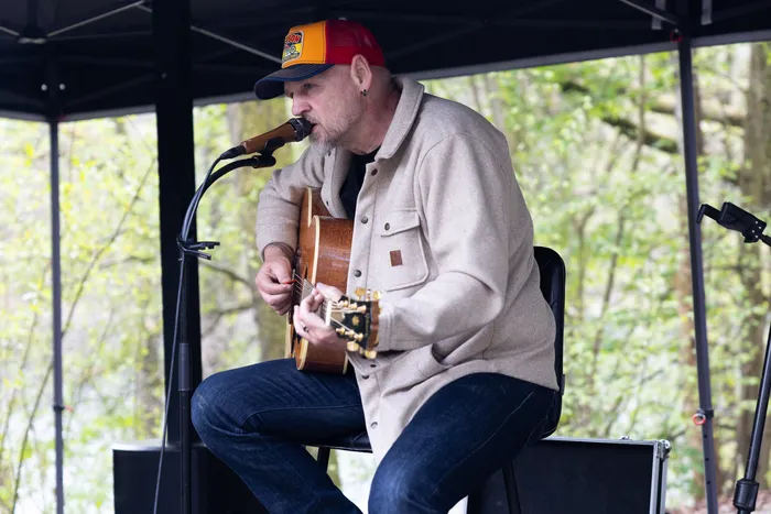 Ein Mann mit roter Baseballkappe spielt Gitarre und singt in ein Mikrofon während einer Veranstaltung im Freien.