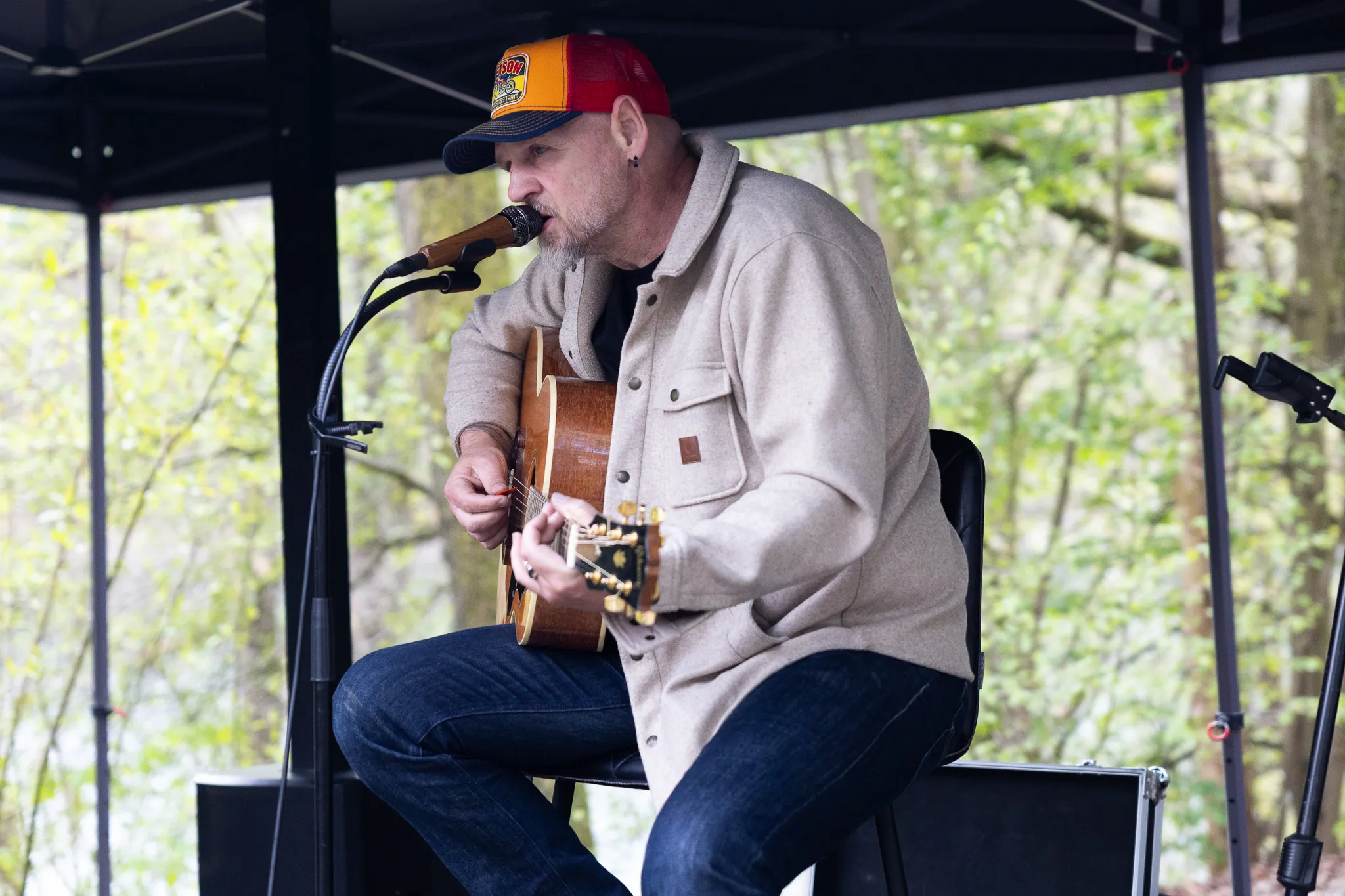 Ein Mann mit roter Baseballkappe spielt Gitarre und singt in ein Mikrofon während einer Veranstaltung im Freien.